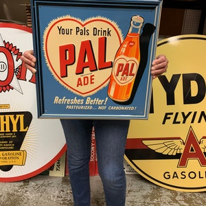 May include: A vintage-style metal sign with a blue background and a heart-shaped design. The sign features the text "Your Pals Drink PAL ADE" and an illustration of a bottle. The sign is framed in black.