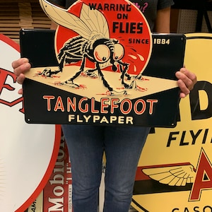 May include: A vintage Tanglefoot Flypaper sign featuring a large fly illustration with the text "WARRING ON FLIES SINCE 1884". The sign is black, red, and cream-colored, with the words "TANGLEFOOT FLYPAPER" in bold red letters.