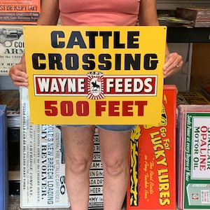 May include: A yellow metal sign with black text that reads "CATTLE CROSSING" and "WAYNE FEEDS 500 FEET." The sign has a red border and a graphic of a cow. Other vintage signs are visible in the background.