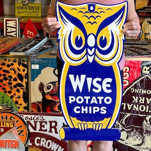 May include: A die-cut metal sign shaped like an owl with yellow, white, and blue colors. The owl features large eyes and a blue chest area with the white text Wise Potato Chips printed in the center. The sign has a small hole at the top for hanging.