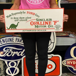 May include: A vintage metal sign advertising Sinclair Opaline "F" Motor Oil. The sign is white with red, green, and black lettering. It features a green dinosaur and the text "Made Expressly for Ford Cars to keep them silent and smooth." and "Authorized Dealer".