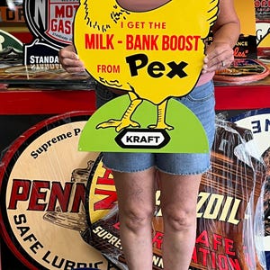 May include: A vintage yellow chicken-shaped sign with black accents and the text "I GET THE MILK-BANK BOOST FROM Pex." The sign has a green base with the Kraft logo. Other vintage signs are in the background.