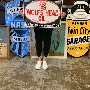 May include: A green and white metal sign with the text "Wolf's Head Oil" in red and white. The sign is surrounded by a white oval with the text "Wolverine - Empire Refining Co." in black. The sign also has the text "A Revelation in Lubrication" in black. The sign is being held by a person wearing a black shirt and black pants.