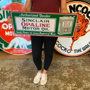 May include: A green and red metal sign advertising Sinclair Opaline Motor Oil. The sign reads "Authorized Dealer Sinclair Opaline Motor Oil Seals Power at every Degree of Wear". A smaller image of a can of Sinclair Opaline Motor Oil is in the upper right corner of the sign.
