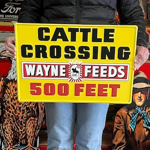 May include: A rectangular yellow metal sign with black text that reads "CATTLE CROSSING" above "WAYNE FEEDS" and "500 FEET" in red. The sign has a logo of a horse inside a circle. The sign is held by a person.