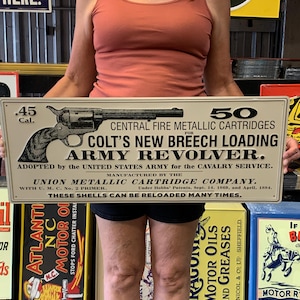 May include: A vintage metal sign advertising Colt's New Breech Loading Army Revolver. The sign features a black and white illustration of the revolver with the text "50 Central Fire Metallic Cartridges for Colt's New Breech Loading Army Revolver. Adopted by the United States Army for the Cavalry Service. Manufactured by the Union Metallic Cartridge Company. With U.M.C. No. 2 Primer. These shells can be reloaded many times."