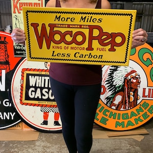 May include: A yellow and black vintage metal sign advertising Woco Pep motor fuel. The sign reads "More Miles", "Trade Mark Reg.", "Woco Pep", "King of Motor Fuel", and "Less Carbon".