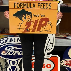 May include: A yellow and orange metal sign with the text "Formula Feeds" and "Feed 'em ZIP". A cartoon pig and bird are running on a yellow track. The sign is held by a person wearing black pants and a pink shirt.