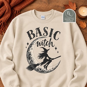 May include: A beige crewneck sweatshirt featuring a black graphic print of a crescent moon and a silhouette of a witch flying on a broomstick. The text "BASIC witch" is printed in black above the moon graphic. The garment has long sleeves.