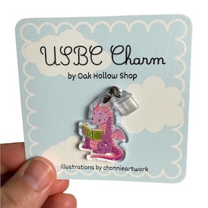 May include: An acrylic charm featuring a purple dragon reading a green book, attached to a light blue display card with white clouds. The card text reads USBC Charm by Oak Hollow Shop and illustrations by chonnieartwork. The charm has glitter.