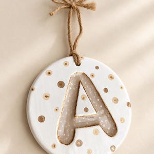 Può includere: Un ornamento circolare bianco da appendere con la lettera A al centro. La lettera è delineata in oro e riempita con un materiale traslucido. La superficie bianca è decorata con piccoli punti dorati e marroni. È fissato un laccio in corda di iuta.
