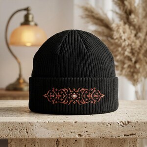 May include: A black ribbed knit beanie featuring a wide folded cuff. Centered on the front of the cuff is an intricate, symmetrical decorative design rendered in a shimmering, glittery copper-toned finish with a small central accent.