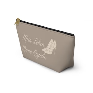 May include: A taupe-colored zippered pouch featuring the white text "Mein Leben. Meine Regeln." and a graphic illustration of two high-heeled shoes. The accessory has a black zipper lining and a gold-toned metal zipper pull at the top corner.