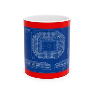 May include: A white ceramic mug with a bright red exterior base and rim. The centre features a blue architectural blueprint graphic of Old Trafford stadium, including technical line drawings, labels, and dimensions in metres for the structure.