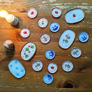 May include: A collection of 14 small, round, and oval ceramic plates with various designs. The plates feature images of ladybugs, mice, and flowers. The plates are arranged on a wooden surface.