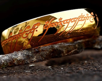 May include: A polished gold-colored ring featuring an engraved script in a dark, reddish-brown color that wraps around the outer surface of the band. The metal surface is highly reflective, showing distorted light reflections.