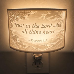 May include: A plug-in night light with a curved, illuminated panel featuring a landscape of mountains and trees. The text "Trust in the Lord with all thine heart ~ Proverbs 3:5" is printed in dark lettering across the center of the glowing surface.