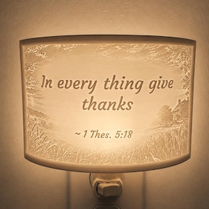 May include: A curved, illuminated night light featuring a textured, embossed landscape scene with a house and wheat field. The centre displays the text In every thing give thanks ~ 1 Thes. 5:18 in a dark, legible font against the glowing white surface.