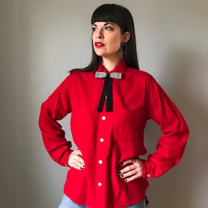 Op de afbeelding: Een rood lang mouw overhemd met knopen en een zwart-wit geblokte strik. Het overhemd heeft een enkele borstzak en wordt gedragen met een blauwe jeans.