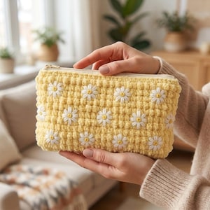 May include: A rectangular yellow crochet pouch featuring a repeating pattern of white daisy flowers with yellow centers. The pouch has a textured, bobble-stitch surface and a light-colored zipper closure along the top edge for secure storage.
