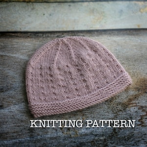 May include: A knitted hat in a light pink color with a textured stitch pattern. The hat has a folded brim.