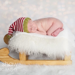 May include: A newborn baby wearing a red and green striped elf hat sleeps on a white fluffy blanket in a wooden sleigh.