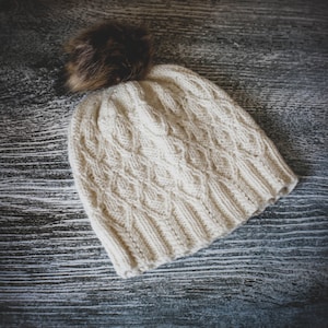 May include: A white knitted beanie with a brown faux fur pom pom. The beanie has a cable knit pattern.