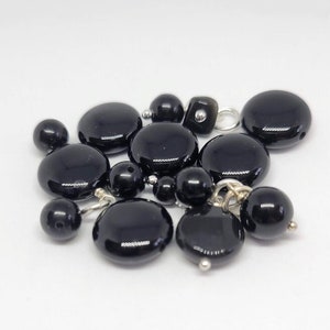 Puede incluir: Una colección de cuentas de ónix negro, algunas redondas y otras planas, con detalles plateados. Las cuentas están dispuestas en un grupo sobre un fondo blanco.