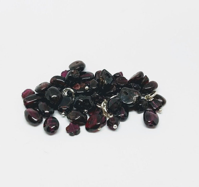 May include: A pile of dark red garnet gemstone beads with silver jump rings. The beads are various shapes and sizes.