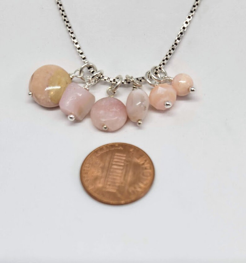 May include: A silver chain necklace with six pink and peach opal gemstone beads. The necklace is about the size of a US penny.