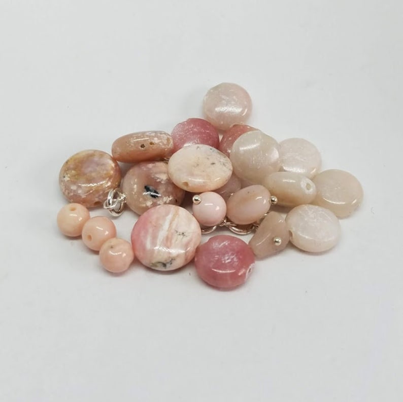 May include: A collection of pink and white opal gemstone beads. The beads are various sizes and shapes, including round, oval, and irregular. Some beads have a striated pattern.