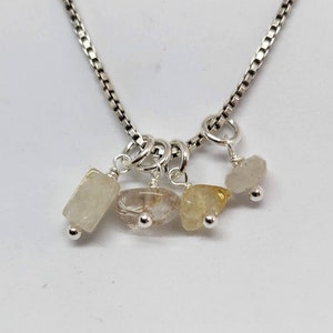 May include: A silver chain necklace with four small, dangling charms. Each charm features a different clear, faceted gemstone: a rectangular white stone, a clear stone with gold flecks, a yellow stone, and a clear, slightly cloudy stone.