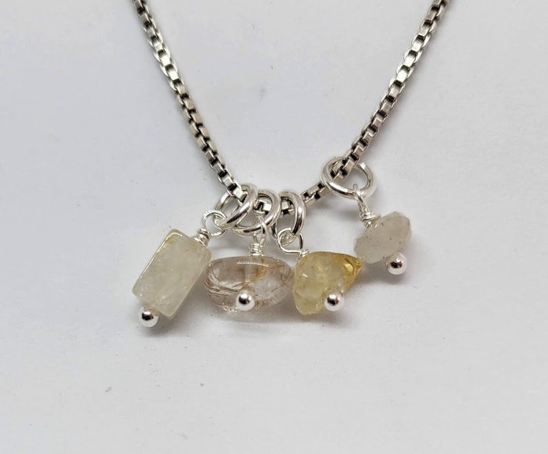 May include: A silver chain necklace with four small, dangling charms. Each charm features a different clear, faceted gemstone: a rectangular white stone, a clear stone with gold flecks, a yellow stone, and a clear, slightly cloudy stone.