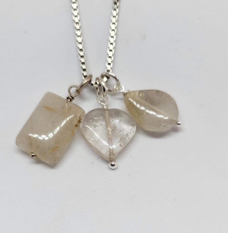May include: A silver chain necklace with three clear quartz crystal pendants: a rectangle, a heart, and a teardrop.