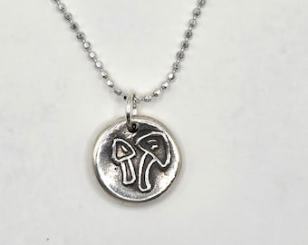 May include: Silver-toned necklace featuring a round pendant with a raised outline of two mushrooms. The pendant hangs from a delicate ball chain. The mushrooms are stylized with curved lines and simple shapes.