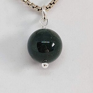 May include: A silver chain necklace with a dark green gemstone pendant. The pendant is a round ball with a smooth, polished surface.