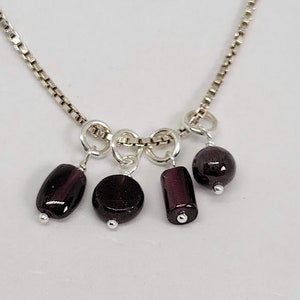 May include: A silver chain necklace with five dangling dark purple gemstone beads. The beads are different shapes, including round, oval, and rectangular.