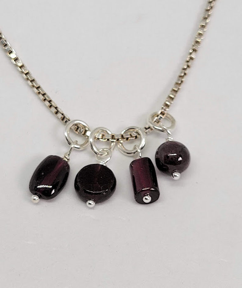 May include: A silver chain necklace with five dangling dark purple gemstone beads. The beads are different shapes, including round, oval, and rectangular.
