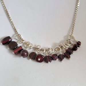 May include: A silver chain necklace with a cluster of dangling garnet gemstones. The necklace features a variety of sizes and shapes of garnet stones, creating a unique and eye-catching design.