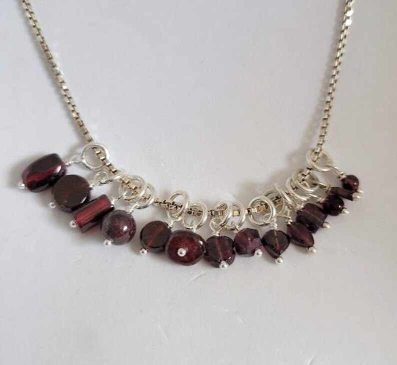 May include: A silver chain necklace with a cluster of dangling garnet gemstones. The necklace features a variety of sizes and shapes of garnet stones, creating a unique and eye-catching design.