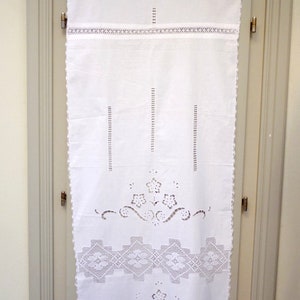 Può includere: Tenda in cotone bianco con dettagli in pizzo intricati e bordi a festoni. La tenda presenta un motivo floreale ripetitivo e un design geometrico.