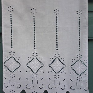 Può includere: Tessuto bianco ricamato con bordo smerlato. Il design presenta file di piccoli fiori, motivi a diamante e linee verticali di piccoli punti. Il tessuto è probabilmente cotone o lino e il ricamo è dettagliato e delicato.