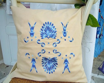 Puede incluir: Una almohada beige con diseños bordados en azul. Los diseños incluyen conchas estilizadas y patrones florales.
