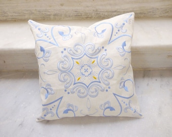 May include: A white pillow with a blue and yellow embroidered design. The design features a central floral motif surrounded by intricate swirls and patterns.