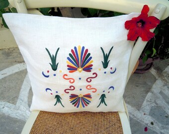Puede incluir: Almohada blanca con diseños bordados de colores. Los diseños incluyen un patrón en forma de abanico con colores rojo, amarillo, azul, verde y naranja, y formas de hojas verdes. La almohada está sentada en una silla de mimbre.