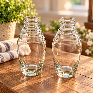 Könnte beinhalten: Zwei durchsichtige Glasvasen mit einer bienenstockartigen Form, die eine breite, abgerundete Basis aufweisen, welche sich nach oben hin in eine Reihe horizontaler, konzentrischer Rippenringe zu einer schmalen Öffnung verjüngt.