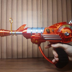 May include: A metallic orange ray gun toy featuring a silver barrel, a black handle, and a circular side dial with the text "Atomic Ray Gun" in gold script. The device includes a small gold circular sight and a lightning bolt emblem on the top.