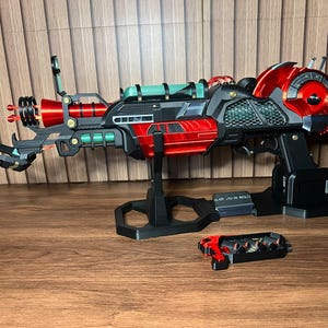Pode incluir: Uma pistola de raios futurista com acabamento metálico vermelho e preto e detalhes em turquesa. O dispositivo inclui um cano com anéis circulares, uma bateria montada na lateral e um suporte. Um pequeno componente separado em vermelho e preto com detalhes internos encontra-se à frente da unidade principal.