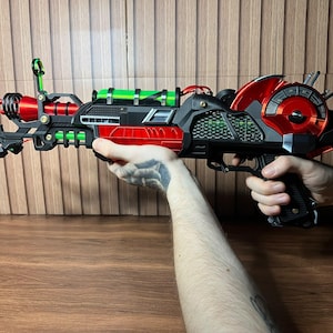 May include: A futuristic ray gun prop featuring a matte black frame with metallic red accents and bright green cylindrical components. The device includes a circular gauge on the side, a front sight, and a textured grip, showcasing intricate mechanical details.