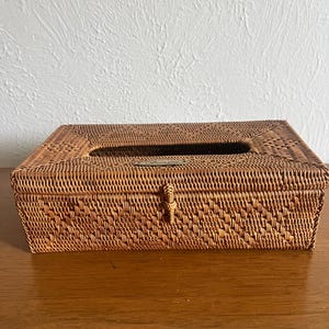 May include: A rectangular tissue box cover crafted from woven brown rattan. The box features a hinged lid with a central rectangular opening for dispensing tissues and a small, decorative metal plaque on the front edge with an engraved logo.